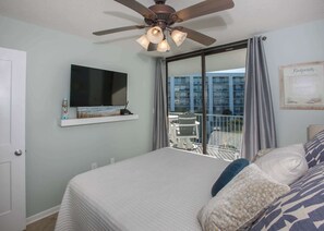 A hotel room with a bed, a ceiling fan, a TV mounted on the wall, and a balcony with a view of a building.
