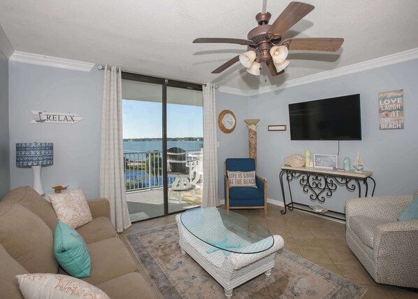 A living room with a large glass table, a blue armchair, and a beige sofa. There is a ceiling fan, a TV, and a view of the ocean through a sliding glass door.