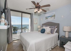 A bedroom with a large bed, a ceiling fan, a nightstand, and a view of the sea through a sliding glass door.