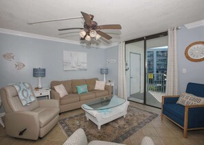Ein Wohnzimmer mit einem beigefarbenen Sofa, einem Glastisch, einem blauen Sessel und einem Deckenventilator.