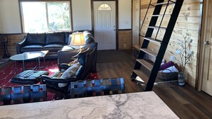 A cozy living room with a leather sofa, a coffee table, and a lamp. There is a wooden staircase with a ladder on the right and a door in the center.