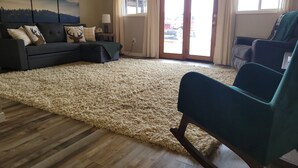 Ein Wohnzimmer mit einer Eckcouch, einem Türkis-farbenen Sessel, einem großen, flauschigen Teppich, Holzboden und einem Gemälde von Bergen an der Wand.