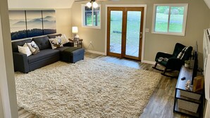 Ein Wohnzimmer mit Ecksofa, großem, flauschigen Teppich, Deckenventilator und Blick durch Glastüren in einen Garten.