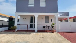 A two-story building with a flat roof, a covered entrance with a door, and a small patio area with chairs and a table. - Sunset Private Studio (Toa Baja)
