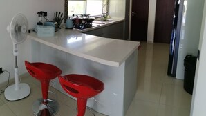 Een moderne keuken met een centraal eiland, rode barstoelen, een staande ventilator en een koelkast.