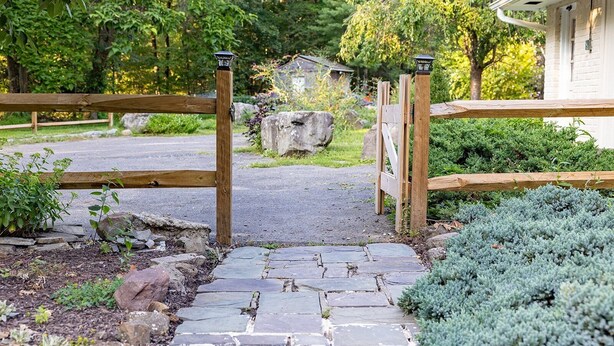 Een houten poort die uitkomt op een oprit met een pad van stenen, omringd door begroeiing en een huis op de achtergrond.