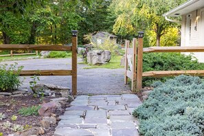 Una verja de madera que da a un camino de entrada con un sendero de piedra, rodeado de vegetación y una casa al fondo.