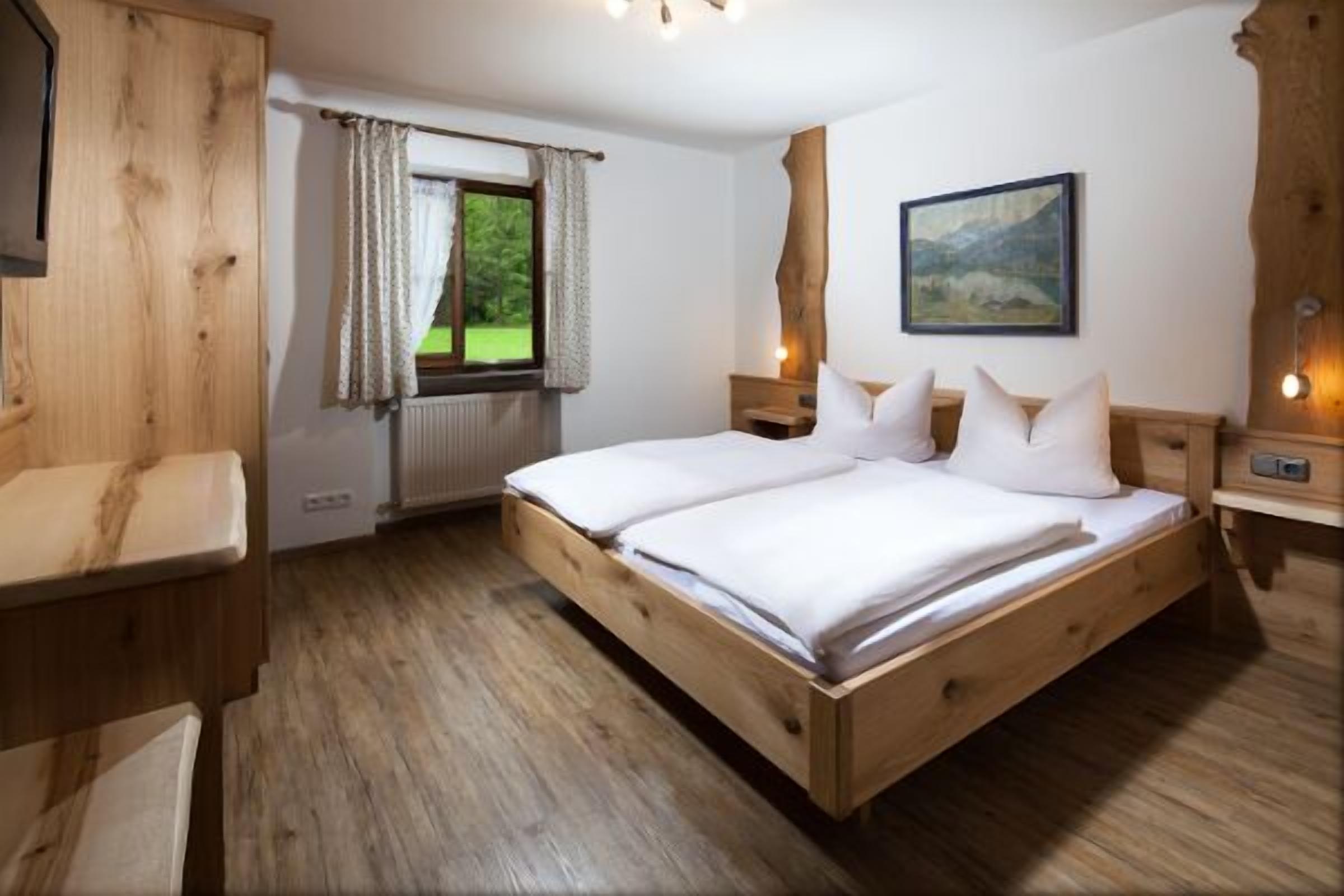 Una cama de madera con ropa de cama blanca, una ventana con cortinas, un cuadro en la pared y una mesita de noche de madera.
