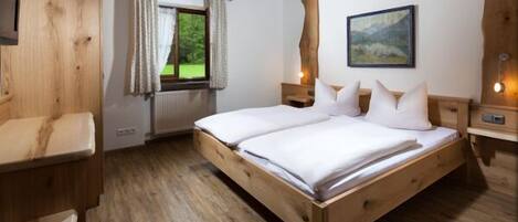 Una cama de madera con ropa de cama blanca, una ventana con cortinas, un cuadro en la pared y una mesita de noche de madera.