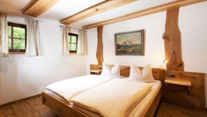 Una cama de madera con colchón y almohadas blancas, una mesita de noche, un cuadro en la pared y una ventana con cortinas.