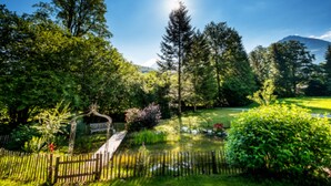 Un jardín tranquilo con un puente de madera, abundante vegetación y una montaña al fondo.