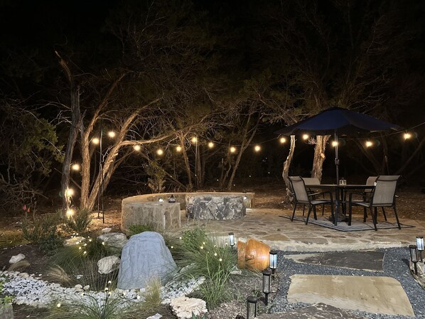 Un espace extérieur aménagé en terrasse, avec une table et des chaises en pierre, entouré d’un éclairage décoratif et comprenant un coin salon abrité.