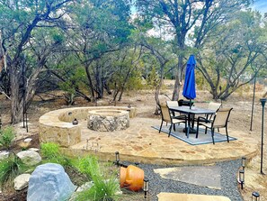 Un foyer en pierre, entouré d’un espace de détente circulaire avec une table et des chaises, est aménagé dans un jardin paysager agrémenté d’arbres et de plantes.