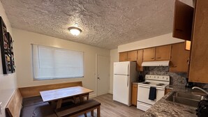 A compact kitchen with wooden cabinets, a white refrigerator, a stove, and a dining table with benches.