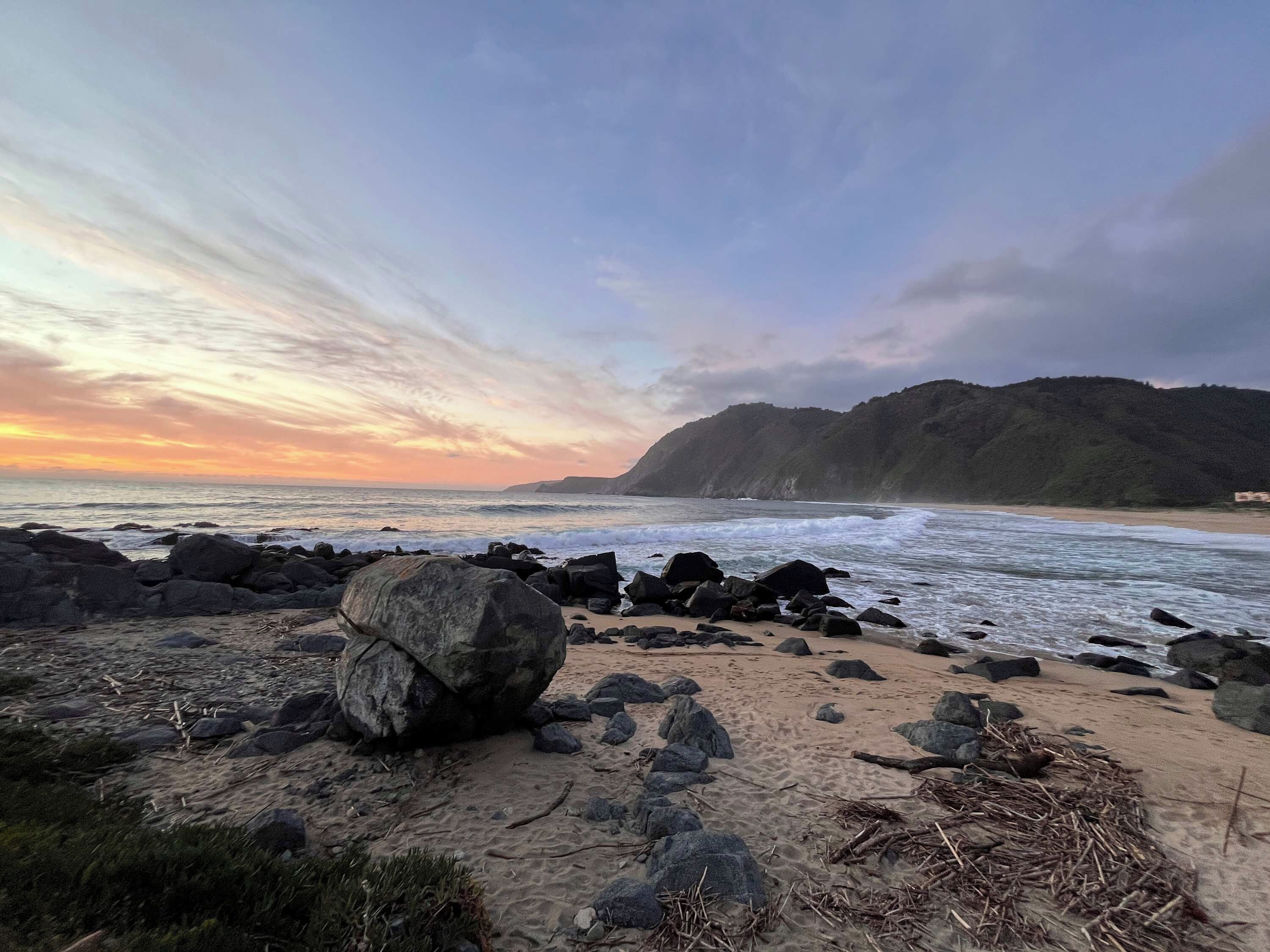 A beach with large rocks in the foreground and a hilly landscape in the background.