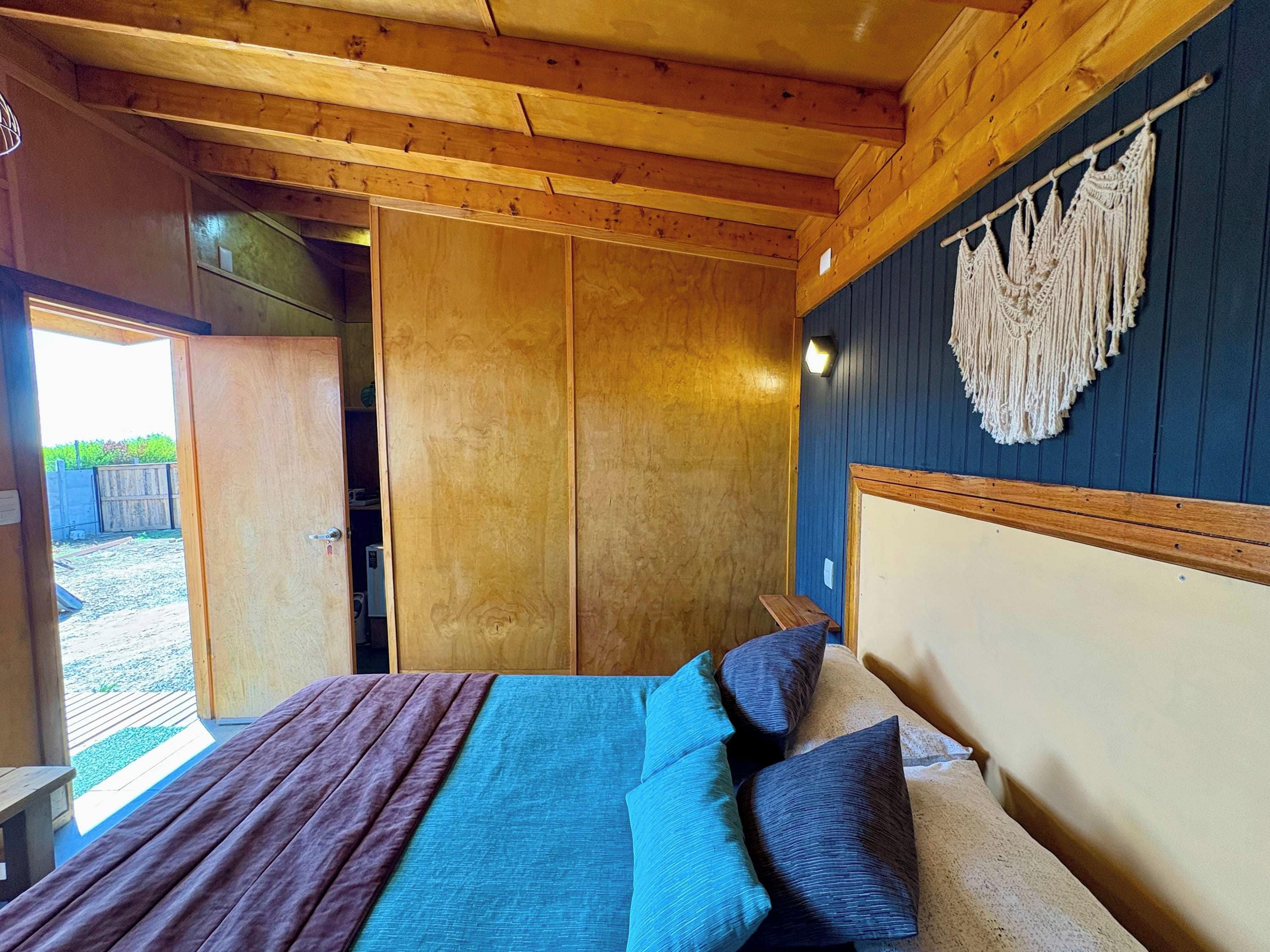 A wooden room with a bed, a wall-mounted macrame piece, and a door leading outside.