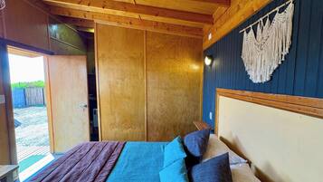 A wooden room with a bed, a wall-mounted macrame piece, and a door leading outside.