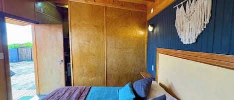 Una habitación de madera con una cama, un tapiz de macramé en la pared y una puerta que da al exterior.