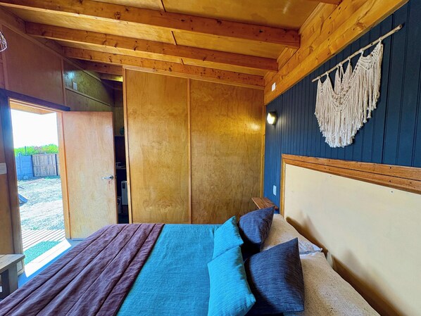 Una habitación de madera con una cama, un tapiz de macramé en la pared y una puerta que da al exterior.