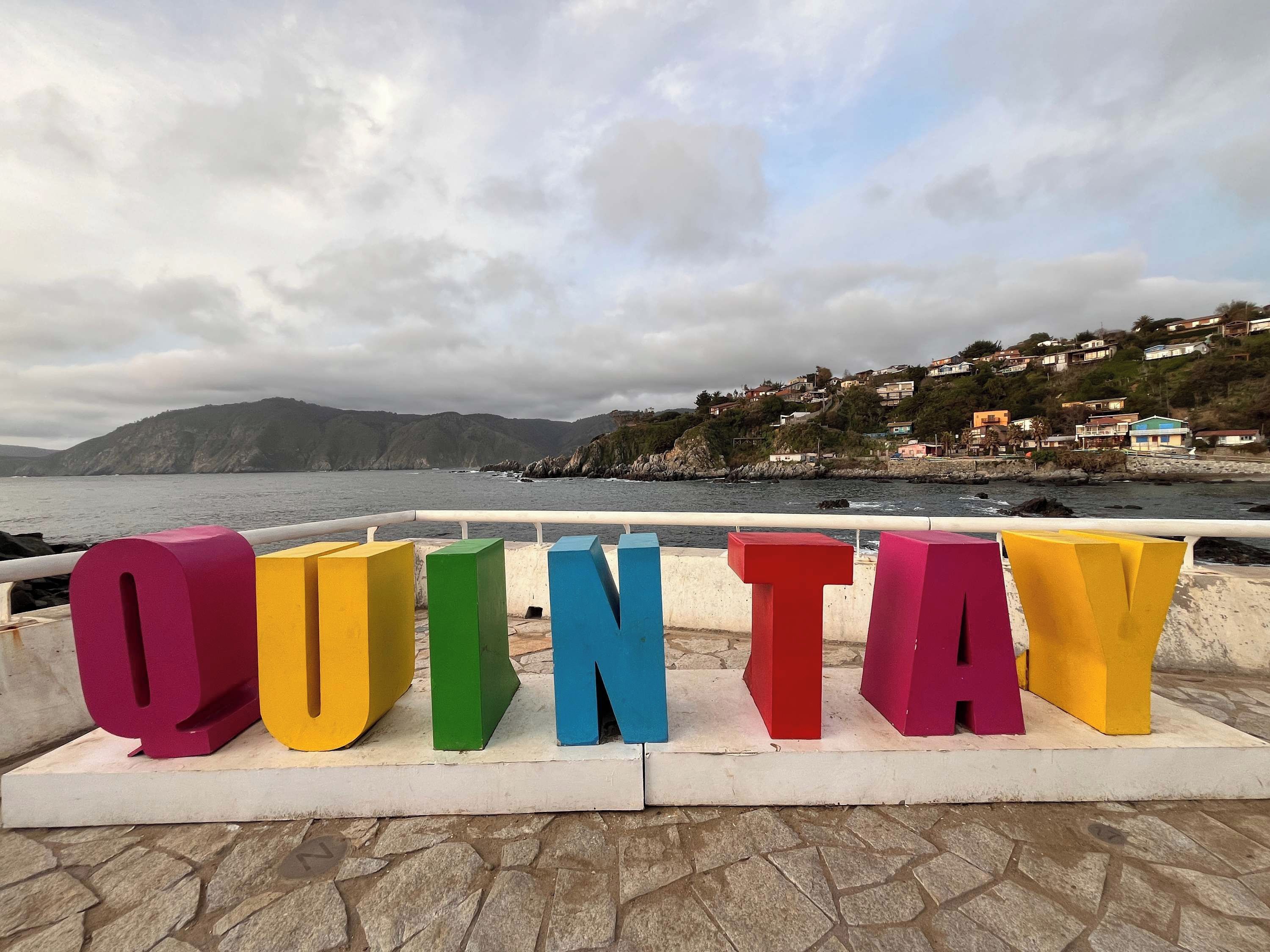 De grandes lettres colorées épellent le mot « QUINTAV » sur une promenade en bord de mer, avec en arrière-plan des maisons et des collines qui bordent la côte.