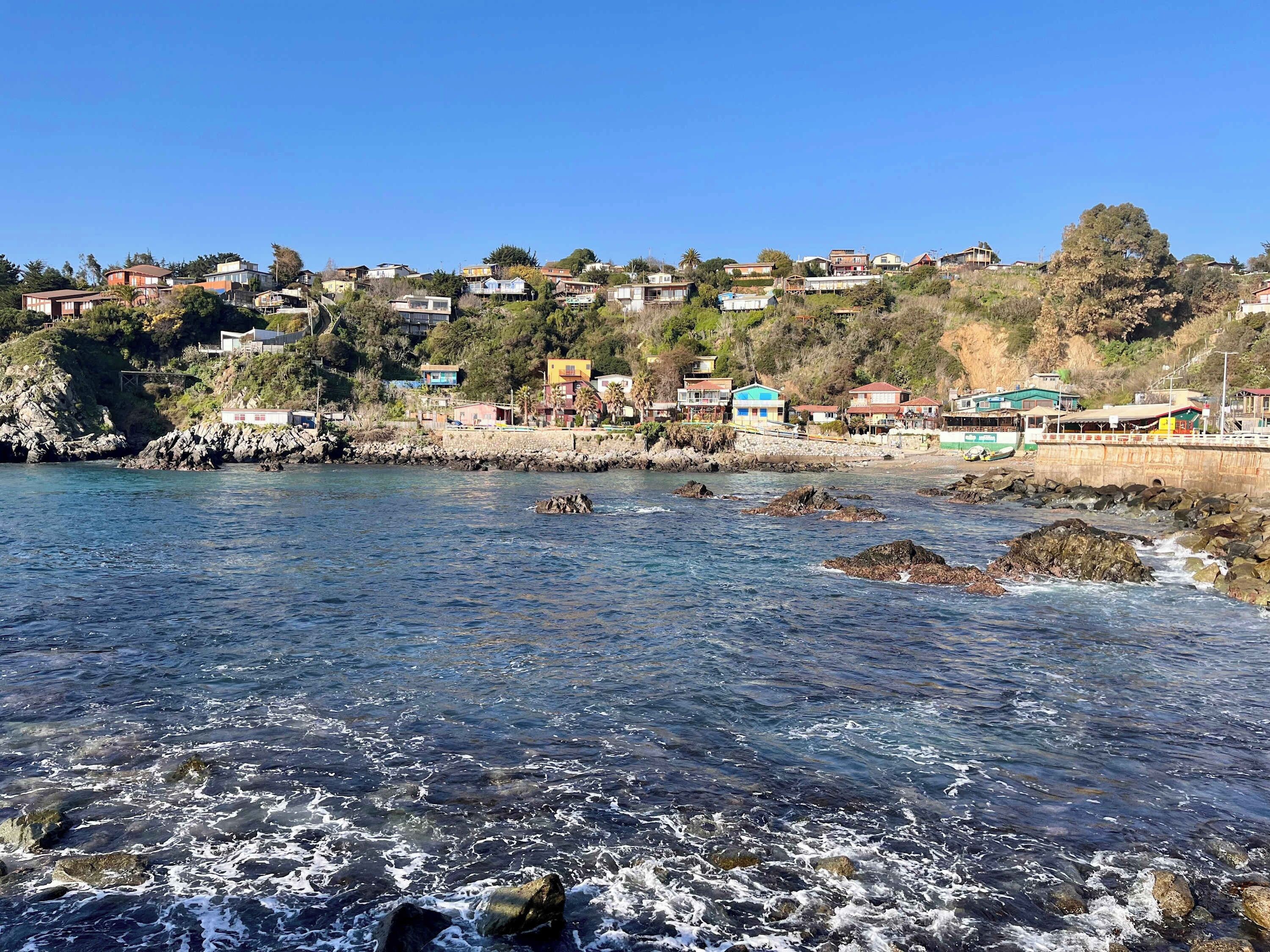 A coastal town with houses on a hillside, rocky shoreline, and clear blue sky.