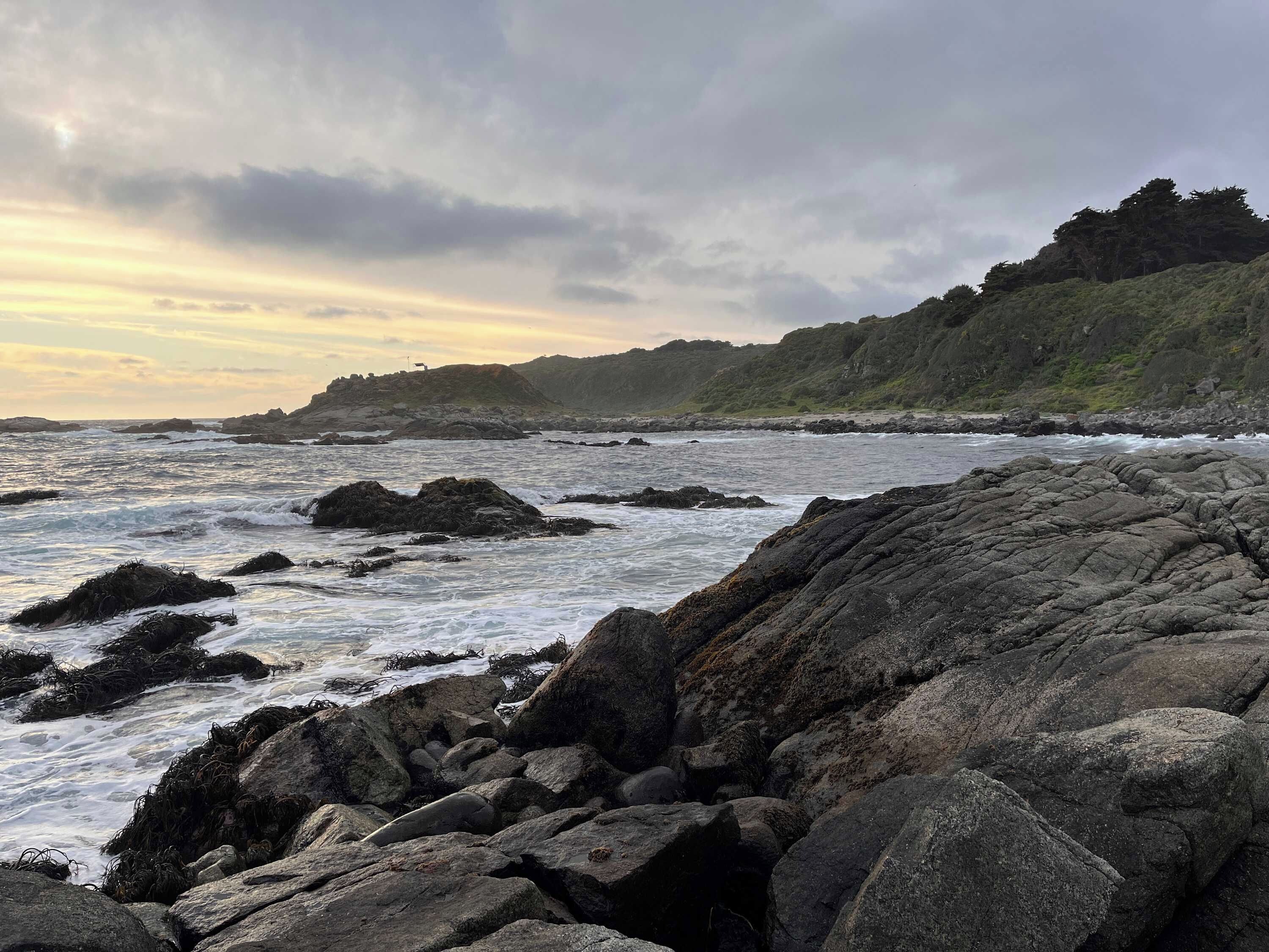 Una costa rocosa con olas rompiendo contra las rocas y una colina al fondo.