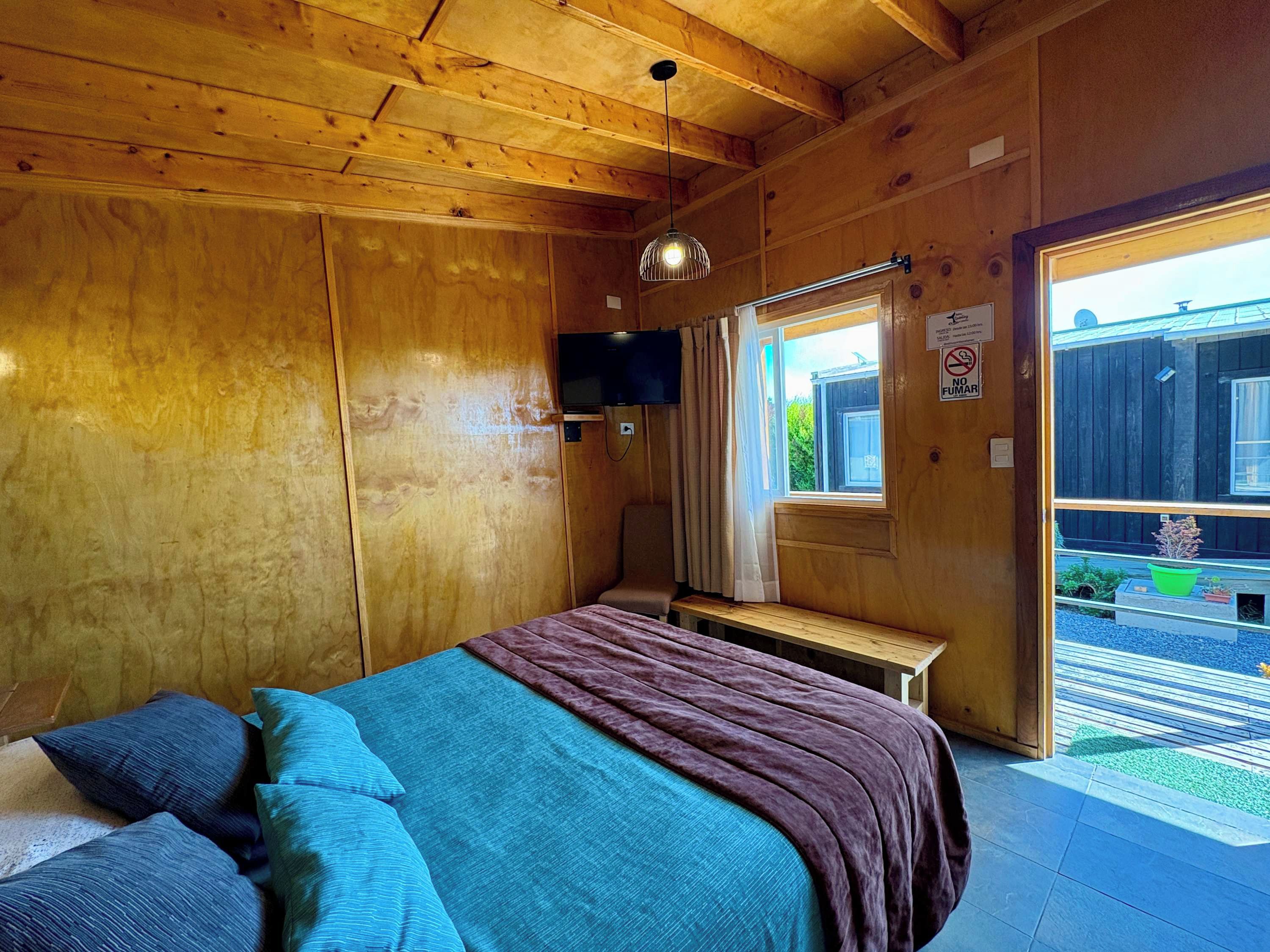 A wooden cabin room with a bed, a bench, a television, and a window with a view of the outside.