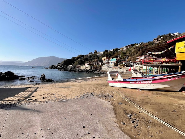 A coastal scene with a boat named 'BAJO MAR' docked on a sandy beach, a paved walkway, and buildings in the background.