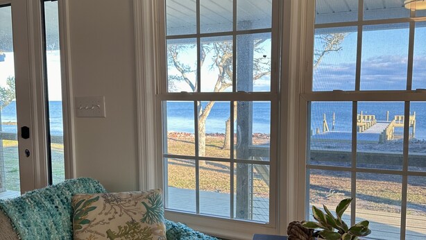 A room with a view of the beach, a couch with a knitted blanket, a table with books and a plant, and a window with a view of the ocean. - Core Sound beauty! Easy access to Core Banks and Cape Lookout. - SLEEPS 11 (Davis)