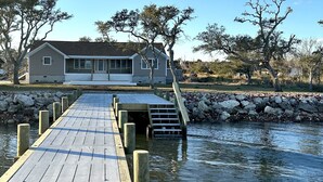 A wooden pier leading to a house by a calm body of water, surrounded by trees and a stone wall. - Core Sound beauty! Easy access to Core Banks and Cape Lookout. - SLEEPS 11 (Davis)