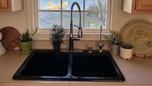 A kitchen with a double sink, wooden cabinets, a window with a view of a building and trees, and several potted plants. - Core Sound beauty! Easy access to Core Banks and Cape Lookout. - SLEEPS 11 (Davis)
