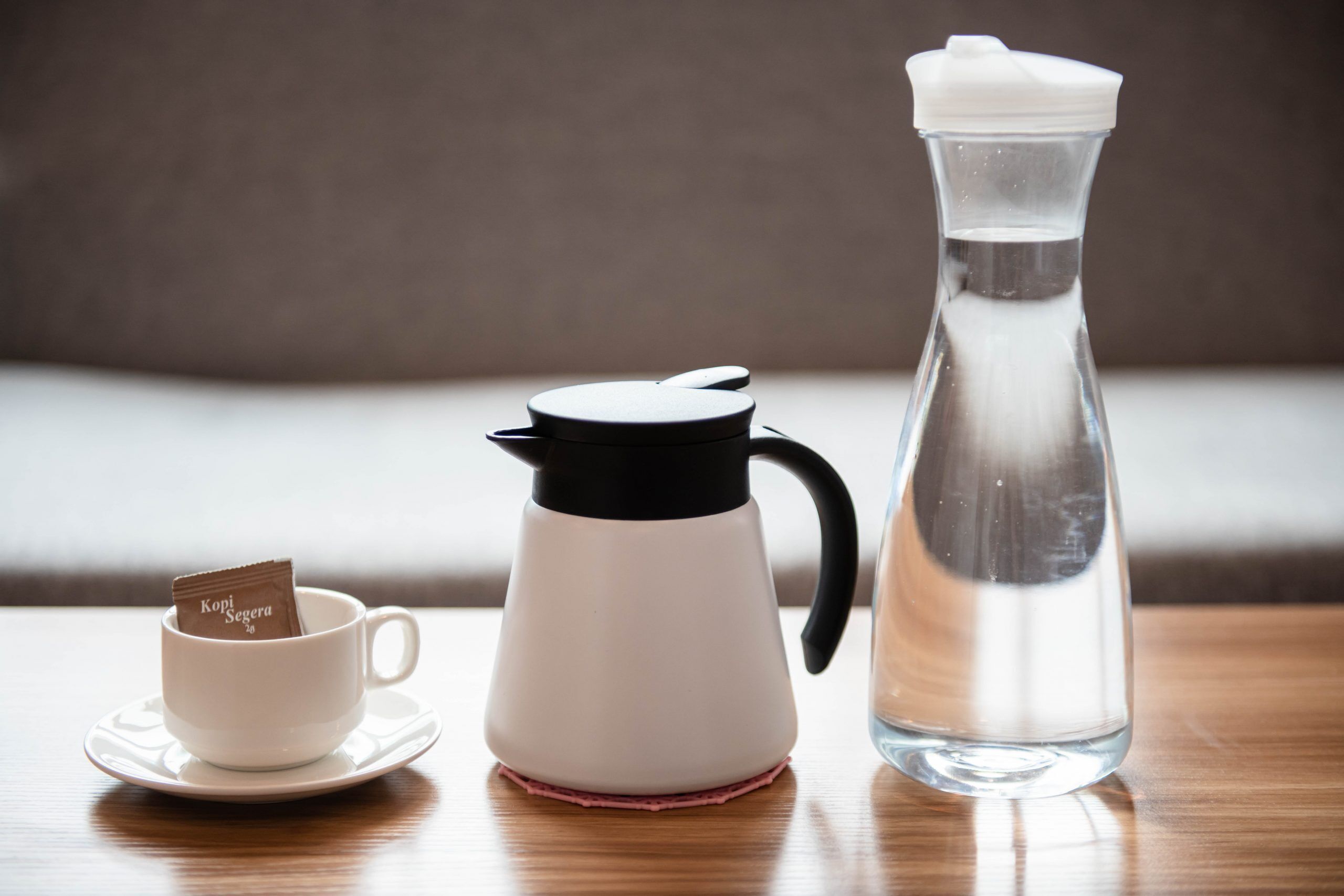 A white coffee maker, a clear water bottle, and a cup with a KOPI coffee sachet on a wooden table.
