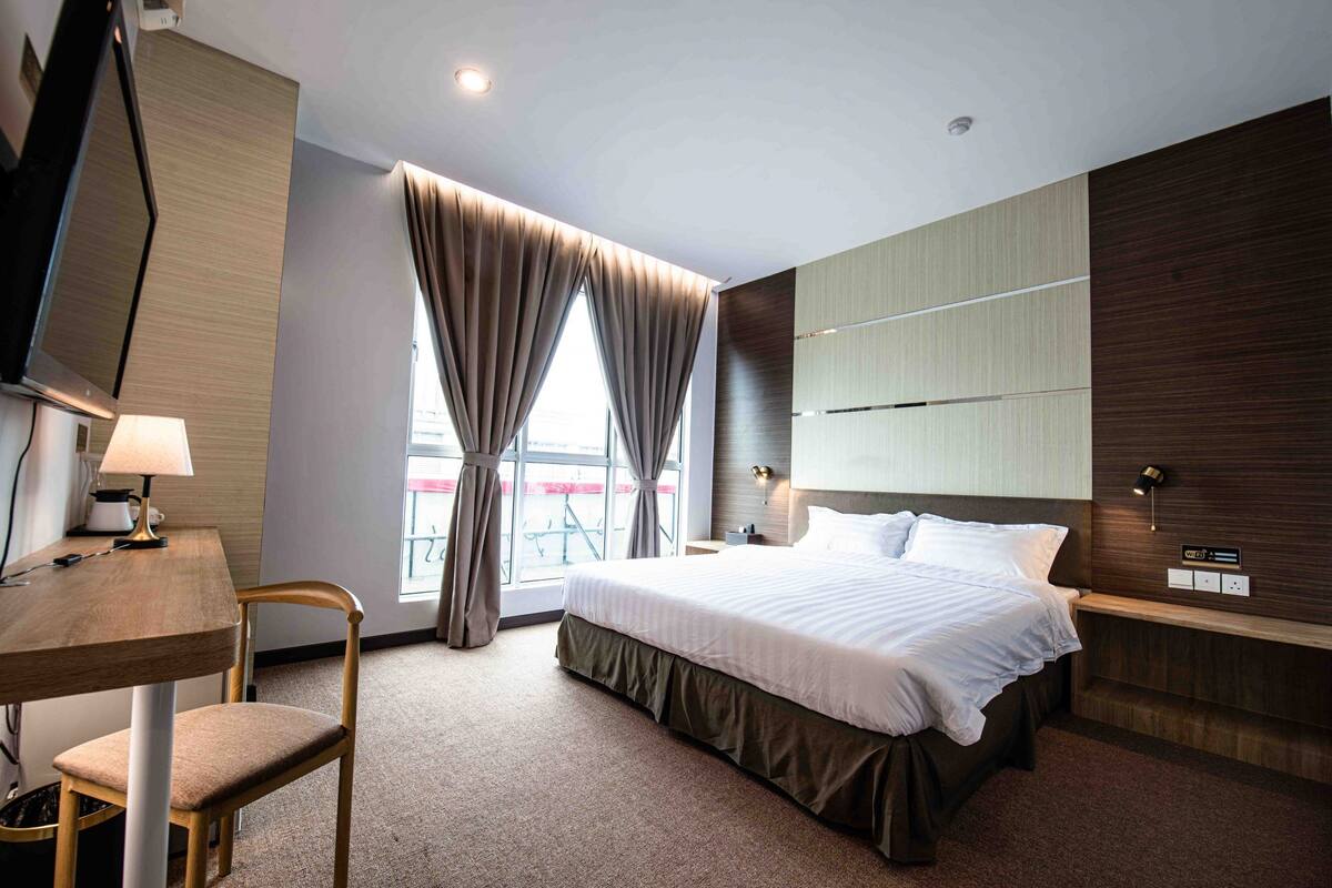 Una habitación de hotel moderna con una cama grande, un escritorio con lámpara, una silla y una ventana con cortinas.