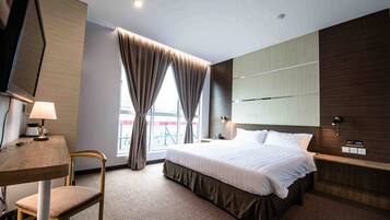 A modern hotel room with a large bed, a desk with a lamp, a chair, and a window with curtains.