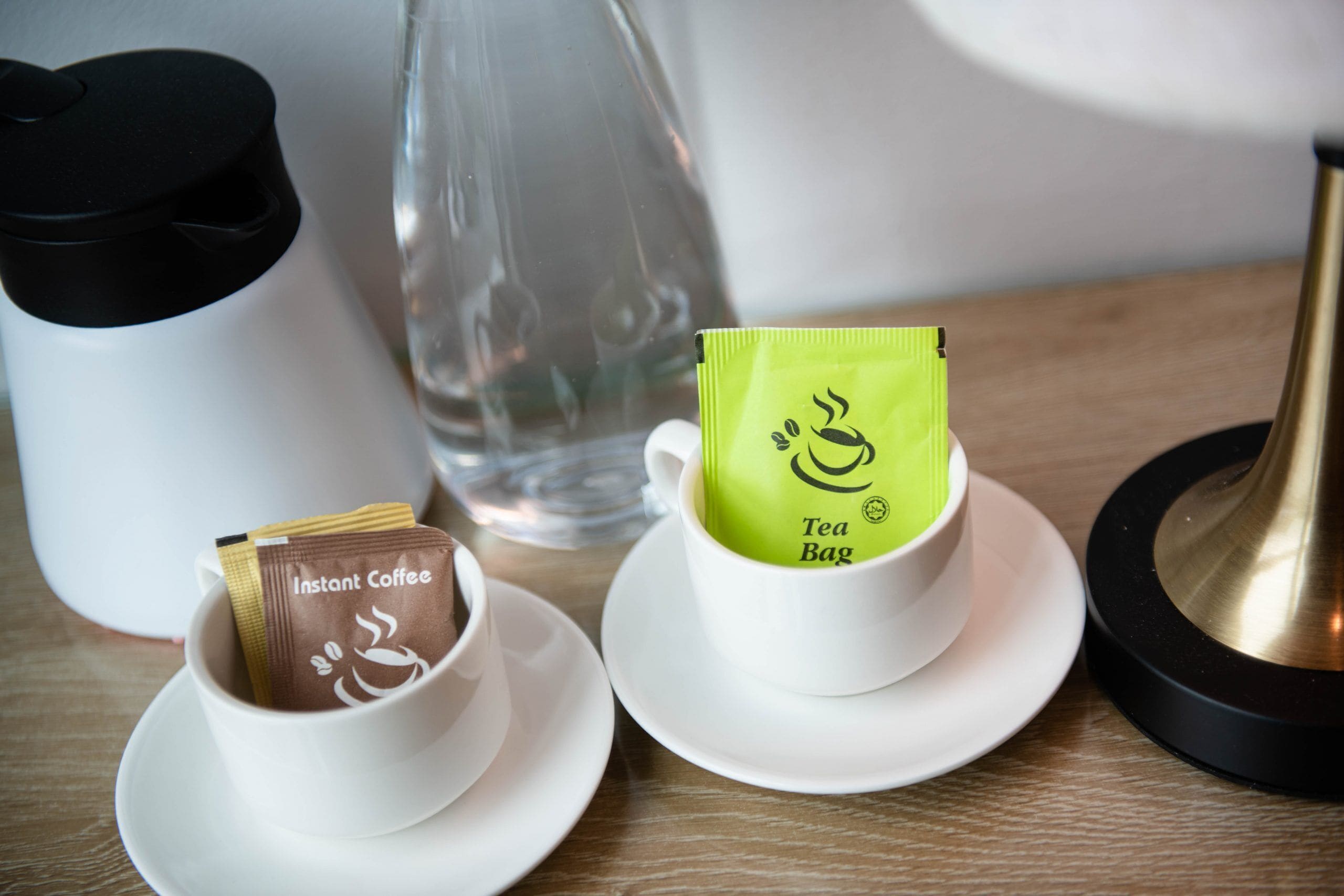 Dos tazas con café instantáneo y bolsitas de té sobre platillos, una botella de vidrio transparente y una lámpara negra y dorada sobre una mesa de madera.