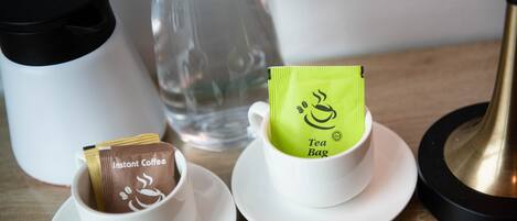 Dos tazas con café instantáneo y bolsitas de té sobre platillos, una botella de vidrio transparente y una lámpara negra y dorada sobre una mesa de madera.