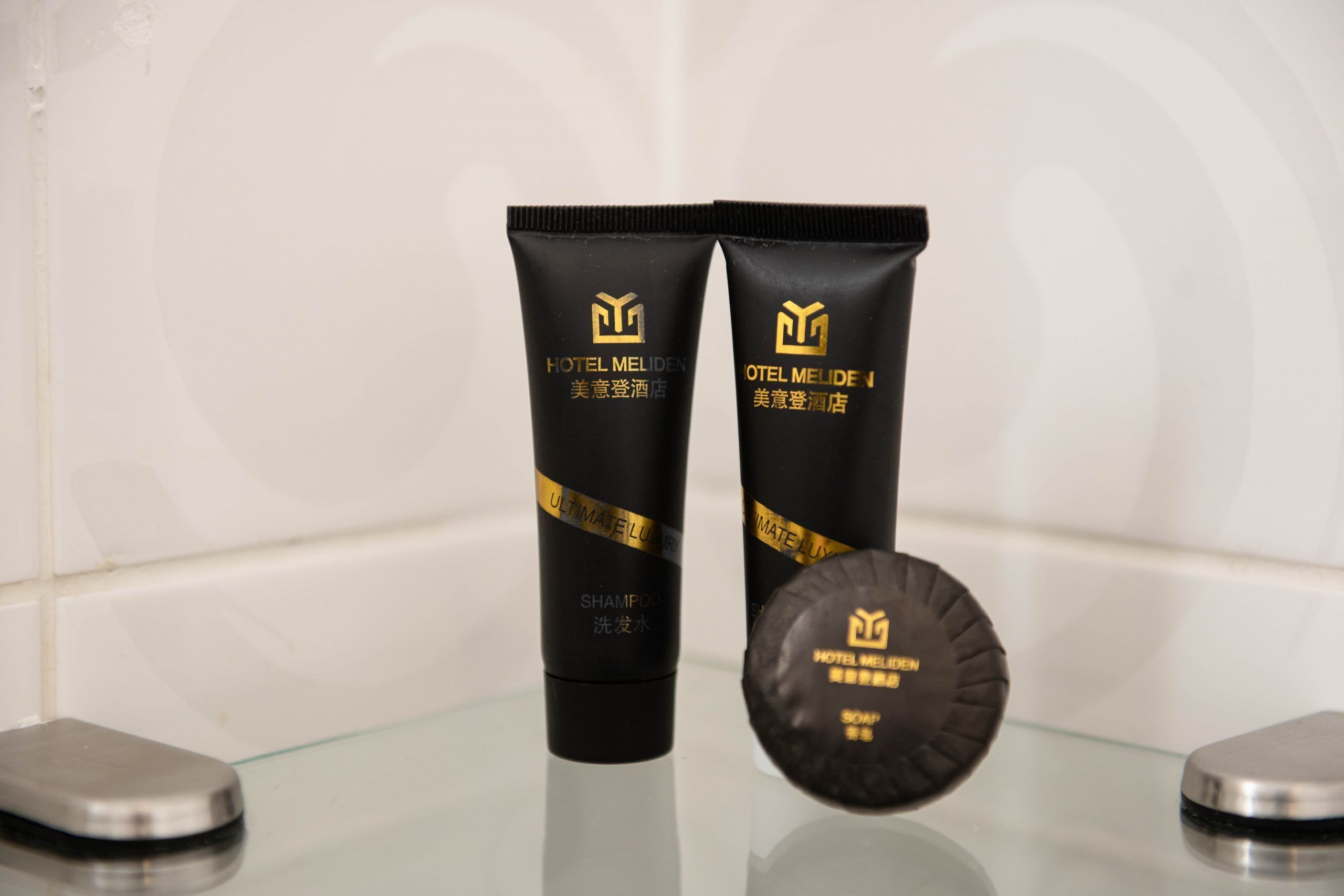 Two black tubes of Hotel Meliden Ultimate Shampoo and a black cap of Hotel Meliden Soap on a reflective surface.
