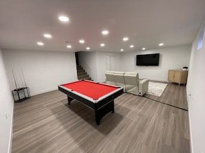 A pool table in a room with a staircase, a flat-screen TV, and a cabinet.