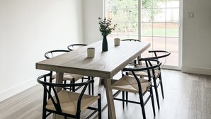 Et spiseområde med et træbord og stole, en vase med blomster, og udsigt til en have gennem en glasdør.