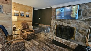 A cozy room with a stone fireplace, patterned chairs, and a television displaying 'RAINY DAY'.