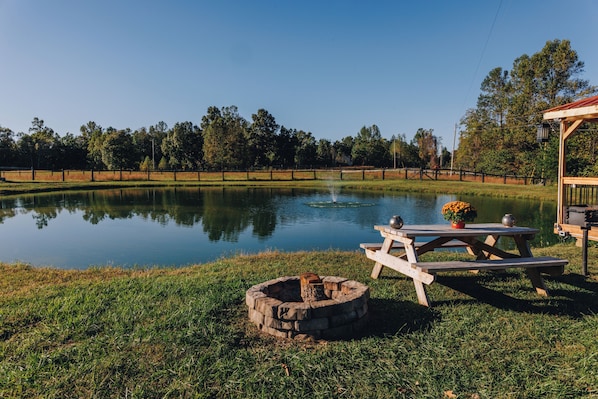 A serene outdoor setting with a wooden picnic table and a fire pit by a calm pond, surrounded by a fence and trees.