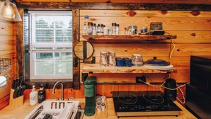 A kitchen with wooden walls, a window, a sink, a stove, and various kitchen items on shelves. - Tiny Cabin At The Pond
 (Stanton)