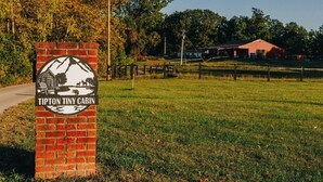 A sign for Tipton Tiny Cabin with a rustic cabin illustration, surrounded by a grassy field and a red barn in the background. - Tiny Cabin At The Pond
 (Stanton)