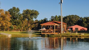 A lakeside cabin with a wooden deck and a red roof, surrounded by trees and a clear sky. - Tiny Cabin At The Pond
 (Stanton)