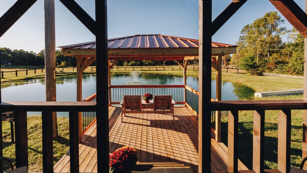A wooden deck with two chairs and a table, overlooking a pond and a metal-roofed structure. - Tiny Cabin At The Pond
 (Stanton)