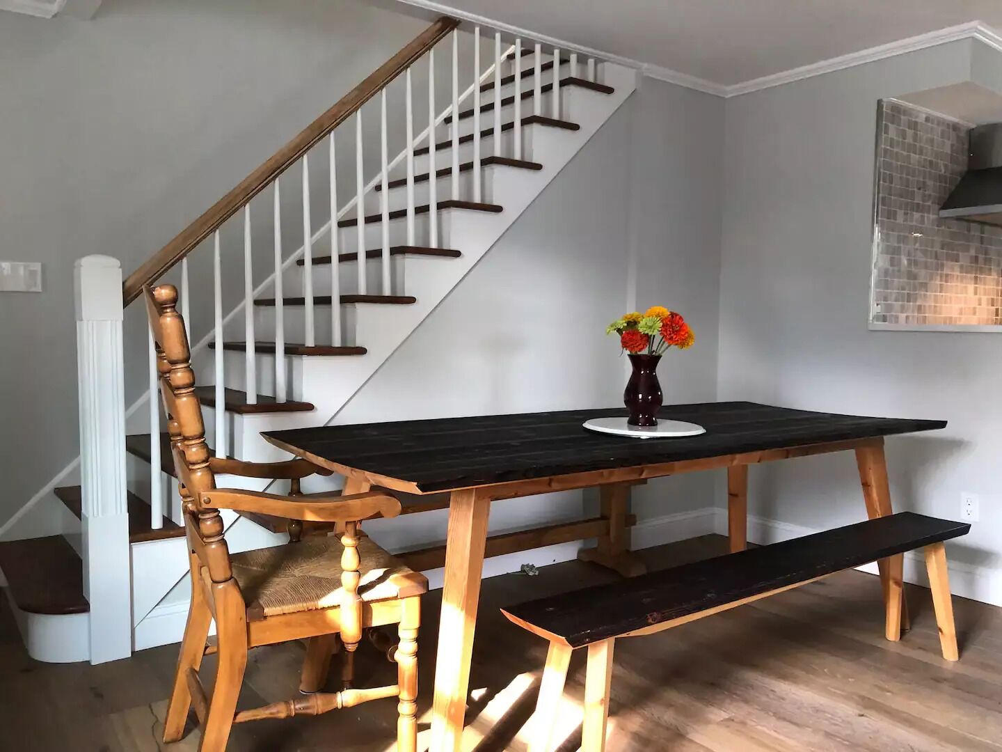 A dining area with a wooden table and bench, a vase with flowers, and a staircase with white railings.