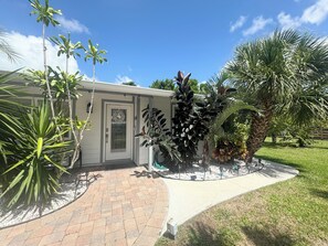 A house with a paved driveway, a front door with a wreath, and various tropical plants.