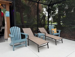 A covered outdoor seating area with Adirondack chairs and a table, surrounded by lush greenery. - Modern Coastal Villa w Luxury Swim Spa & Art (Sarasota)