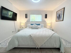 A bedroom with a large bed, two bedside tables, a television mounted on the wall, and a window with blinds. - Modern Coastal Villa w Luxury Swim Spa & Art (Sarasota)