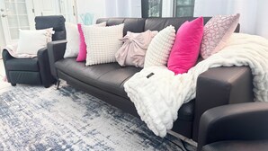 Ein Wohnzimmer mit einem dunkelbraunen Sofa, mehreren Kissen in verschiedenen Farben, einer weißen, flauschigen Decke und einem gemusterten Teppich.
